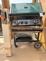 Full view of Napoleon gas grill cart with tools on shelf and boxed Weber rotisserie leaning against side