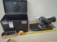Full lot showing case, sewing machine on board, tape measure for scale, box of attachments