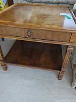 Front view of medium brown veneer end table showing drawer with brass ring pull and open shelf