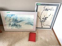 Both framed prints leaning against wall with red Chinese Art book on carpet