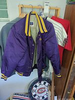 Front view of purple varsity jacket on hanger with towel, dartboard and bat partially visible