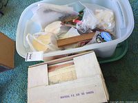 Plastic tote containing yarn rolls, canvases, pattern and accessories alongside cardboard box with printed canvas