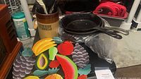 Lot overview showing crock with utensils, three cast iron skillets, silicone lids and metal fruit wall art
