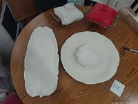 Overview of five ceramic pieces on table: leaf platter, round platter with bowl, red and white lidded casseroles on metal stands