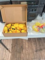 Box containing numerous yellow rubber ducks with sunglasses next to bagged assorted ducks