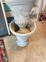 Two matching grey plastic urn planters, one nested inside the other showing decorative details and pedestal bases