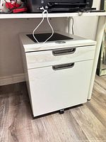 Front view of white two-drawer mobile file pedestal under desk showing handles, lock, and caster