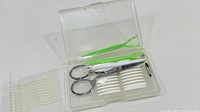 Plastic case open showing scissors, two green grooming tools, black pin tool, and translucent curved plastic pieces; separate tray of identical curved pieces nearby
