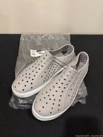 Pair of grey perforated slip-on shoes on plastic packaging, angled view