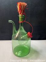 Front view of green glass decanter showing side spout, raffia stopper, hanging small reservoir