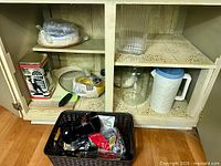 Open cupboard showing glass jar, pitcher, tin, cutting boards, plates, basket