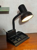 Front-right view of black gooseneck desk lamp illuminated, showing organizer compartments
