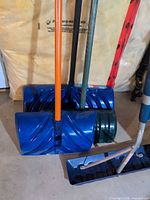 Three upright snow shovels and roof rake lying on floor