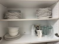 Two cabinet shelves showing stacks of white irregular-edge plates, decorative bowls, mugs and clear glasses