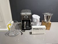 Group shot of all five items: coffee maker, Brita pitcher, filter box, glass pitcher, stainless bucket