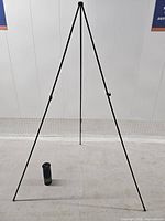 Assembled black tripod easel with small carry piece on floor for scale