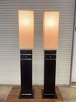 Front view of both black column floor lamps with lit off-white shades