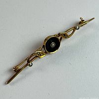 Oblique view of entire brooch showing black enamel center, seed pearls, gold scrollwork and C-clasp