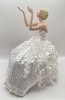 Full left profile of seated porcelain doll in white gown, arms raised