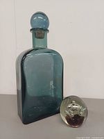 Teal glass decanter with stopper alongside clear art glass paperweight