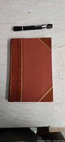 Front cover of brown leather hardcover with gilt decoration next to pen for scale
