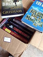 Box containing multiple hardcover books showing The Broker and Weapons Of Your Warfare on top