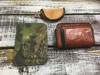 Three small accessories on rustic wood surface: tan half-moon leather coin purse, camo card holder, reddish-brown zip coin purse