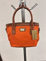 Front view showing orange nylon body, brown handles, gold-tone logo plaque, attached hangtag