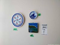Wall display showing 6.5 in floral trivet, 3 in sailing ship plate, 4x4 in landscape tile