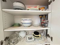 Cupboard shelves showing entire assortment: Saga plates, teapots, scale, bowls, dishes, battery packs, goblets