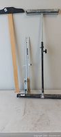 Three tools upright: wood-handle squeegee, aluminum measuring guide, steel floor roller
