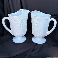 Two white milk glass pitchers side by side showing embossed pattern and pedestal bases