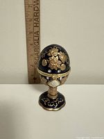 Front view of blue enamel egg trinket box on pedestal with ruler for scale