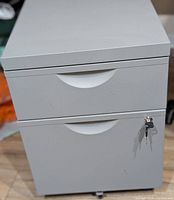 Front view of grey 2-drawer steel filing cabinet showing recessed pulls, lock, and keys inserted, cabinet on casters