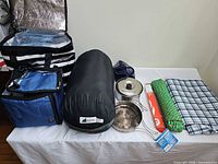Full lot view on table showing sleeping bag, two cooler bags, cookware, rope, tablecloth, matches