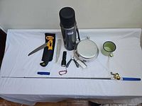 All items laid out on table including fishing rod, thermos, mess kit, mug, saw, flashlight, multi-tool, fire starter