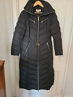 Front view of black quilted long coat on hanger showing hood, belt, gold zip hardware