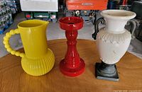three decor items on table: yellow pitcher, red candle holder, white vase