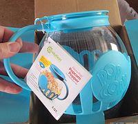 Popcorn popper in box showing glass bowl, turquoise silicone handle and band