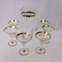 Full view of cocktail set showing one pitcher with handle and six glasses, all clear glass with gold rims on the top and bottom edges.
