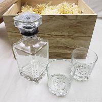 Chunky clear glass decanter with stopper and two highball glasses placed in front of the wooden crate filled with natural shredded paper filler.