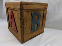 Three photos showing various sides of a wooden cube toy box/stool. Each side has a raised alphabet letter 'A', 'B', or 'C' painted in different colors (red, blue, white). The box has a natural wood finish and a hinged lid top, about 11 1/2 inches tall and 11 inches wide.