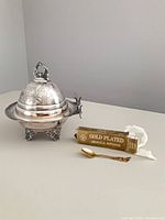 Silver-plated butter dish with dome lid beside boxed gold-plated spoon