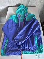 Full view of Columbia purple and teal windbreaker laid flat showing front, hood, pockets, drawstring hem