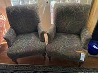 Pair of matching floral tapestry armchairs side by side on hardwood floor, turned legs visible, auction tag attached