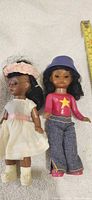 Front view of both dolls showing outfits, accessories and measuring tape for scale
