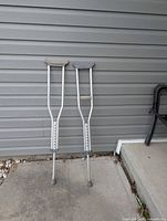 Full view of both aluminum crutches standing upright against siding