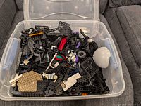 Top view of clear plastic bin filled with assorted black and colored LEGO parts; visible white dome, tan plate, various vehicle pieces