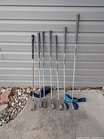 Full set of seven visible clubs leaning against wall with two headcovers on ground