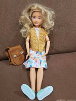 Doll wearing yellow floral shirt, patterned skirt, teal sneakers; brown backpack adjacent